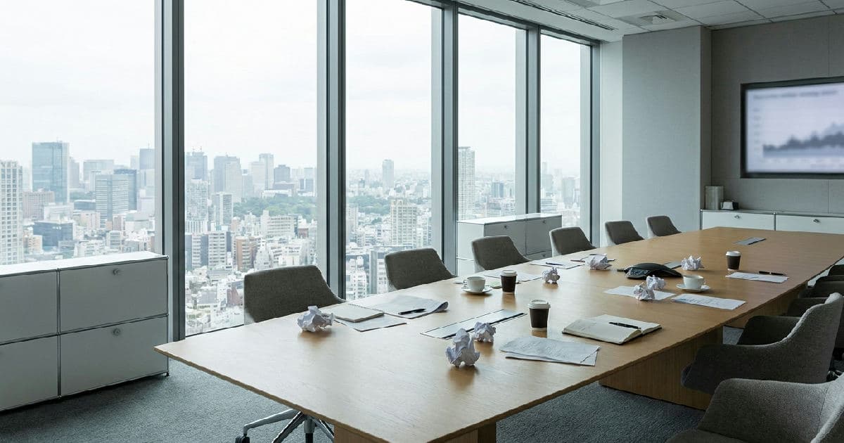 Empty boardroom with meeting table, papers, and city views through windows.